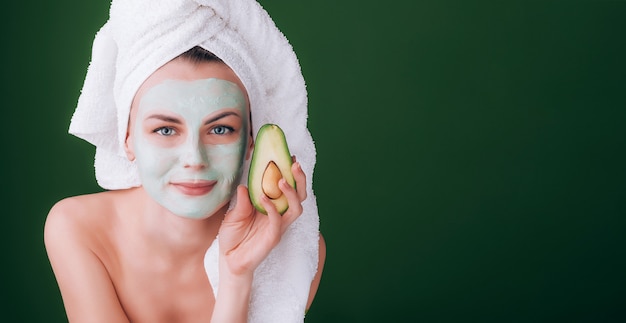 Meisje met een witte handdoek op haar hoofd met een voedzaam groen masker op haar gezicht en een avocado in haar handen op een groene achtergrond met ruimte voor tekst