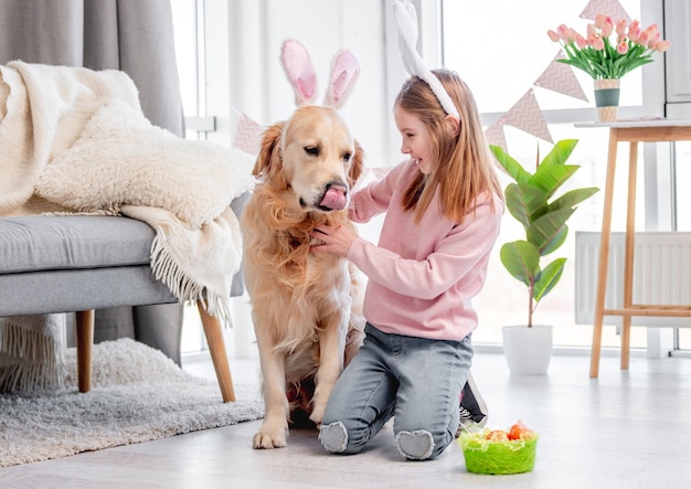 Meisje met gouden retrieverhond die konijntjesoren dragen die op de vloer thuis op Paasdag zitten