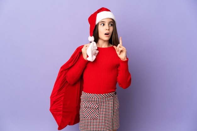 Meisje met hoed en Kerstmiszak die op purpere muur wordt geïsoleerdm die een idee denken dat de vinger omhoog wijst