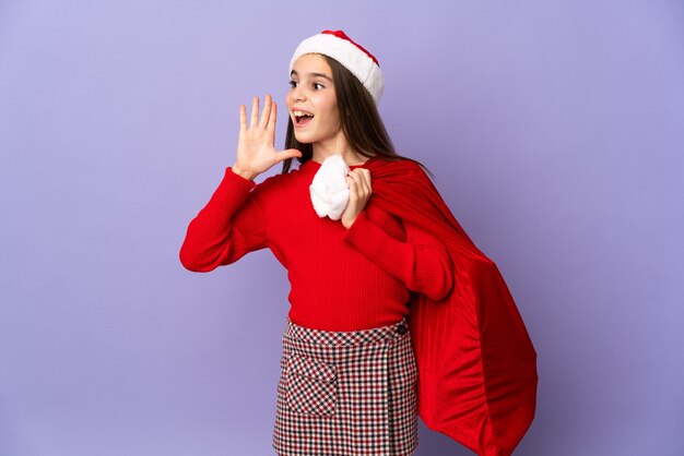 Meisje met hoed en kerstzak geïsoleerd op paarse muur schreeuwen met mond wijd open aan de zijkant