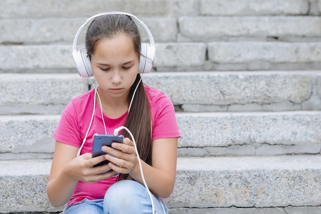 Meisje met koptelefoon zit op de trap met een smartphone in haar handen. Lang haar, roze t-shirt, blauwe spijkerbroek.