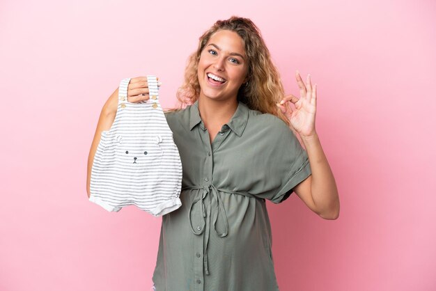 Meisje met krullend haar geïsoleerd op roze achtergrond zwanger en houden babykleding met ok teken