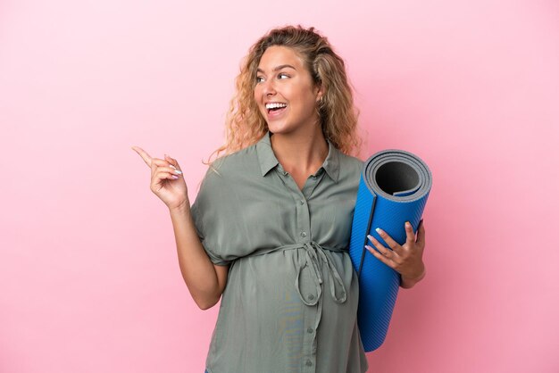Meisje met krullend haar geïsoleerd op roze achtergrond zwanger en houdt een mat vast terwijl ze iets presenteert