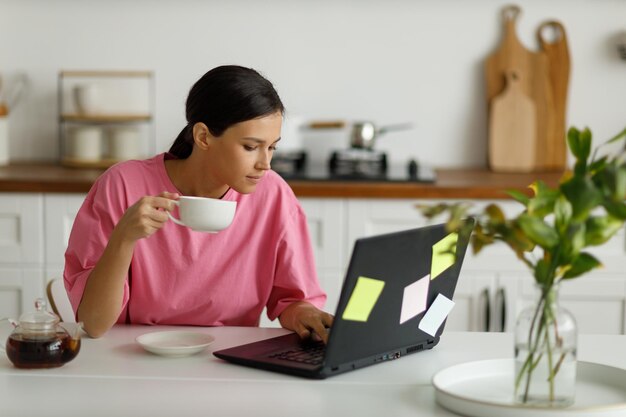 Meisje met laptop controleert haar mail, kijkt door berichten of grappige video's Videocommunicatie online Jonge vrouw werkt op afstand vanuit huis zittend aan de eettafel in de keuken