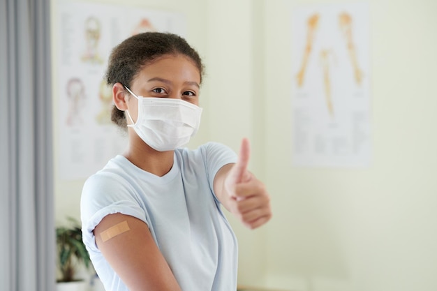 Meisje met masker gebaren met duim omhoog en laten zien dat ze ok is na vaccinatie terwijl ze in het ziekenhuis staat