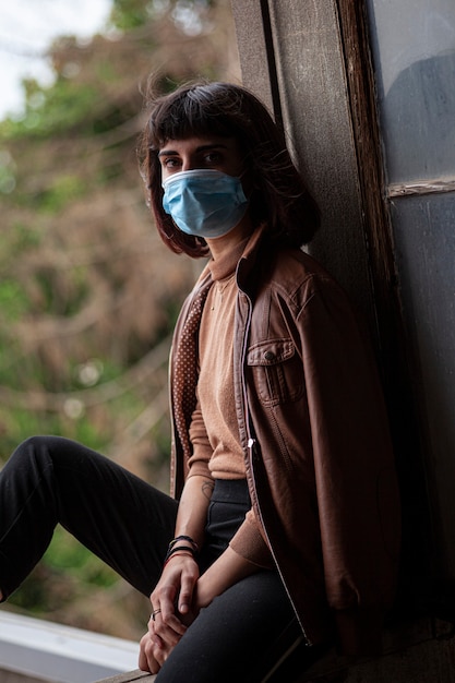 Meisje met medisch masker bij raam in haar huis tijdens covid quarantaineperiode