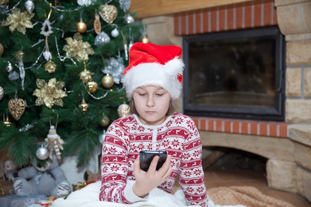 Meisje met mobiele telefoon bij kerstboom in woonkamer