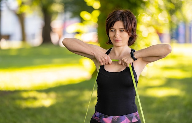 Meisje met rubberen elastische band die armweerstandstraining buiten in het park doet Jonge vrouw die 's ochtends traint met extra apparatuur