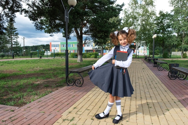 Meisje met rugzak schooluniform met witte strikken en stapel boeken in de buurt van school Terug naar school gelukkige leerling zware leerboeken Onderwijs basisschoolklassen 1 september