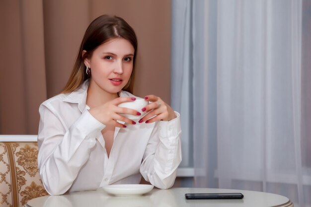 Meisje met Slavische verschijning met telefoon en witte kop zit aan tafel