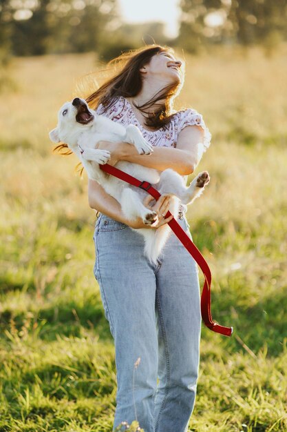 Meisje met speelse schattige pluizige puppy hilarisch moment Spelen met hondje Gelukkige jonge vrouw met grappige witte puppy in warm zonsonderganglicht in de zomerweide