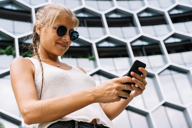 Meisje met zonnebril die zich voor een modern gebouw bevinden die met mobiel texting