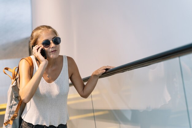 Meisje met zonnebril en een zak die van haar schouder hangt die op haar mobiele telefoon in een roltrap spreekt