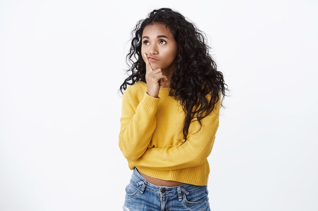 Meisje nadenken over ideeën. nadenkend dwaas en schattig afro-amerikaans meisje in gele trui, kijkend in de linkerbovenhoek, lip aanraken als denken, inspiratie zoeken, plan in gedachten hebben, witte muur