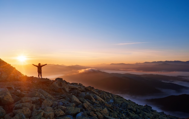 Meisje op een bergtop met uitgestrekte armen. Dramatisch landschap van een eenzame wandelaar die bergen in mist bij zonsopgang bekijkt. Vrijheid, actieve levensstijl en overwinningsconcept.