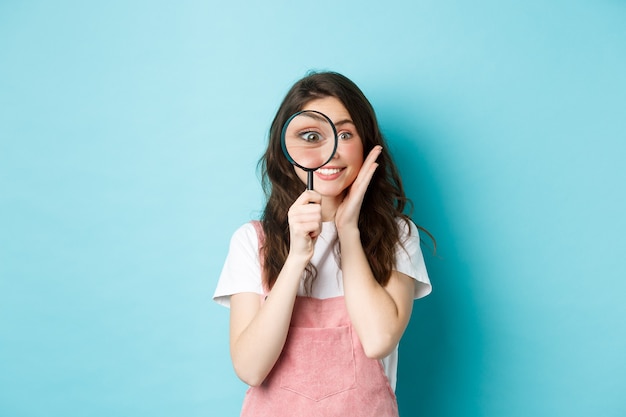 Foto meisje op zoek naar jou. schattige lachende vrouw recruiter kijkt door vergrootglas, starend naar de camera, onderzoekend, op zoek naar iemand, staande over blauwe achtergrond