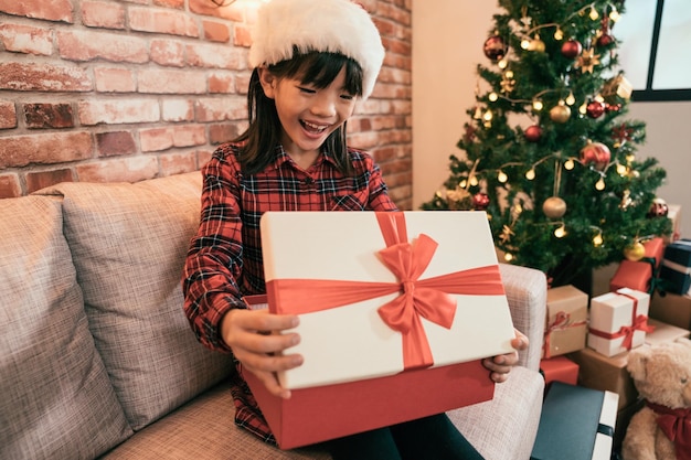 meisje opgewonden open het geschenk van de ouders. klein kind zittend op de bank alleen thuis op de tweede kerstdag. gelukkig kind met kerstmuts die vrolijk lacht in de gezellige kamer.