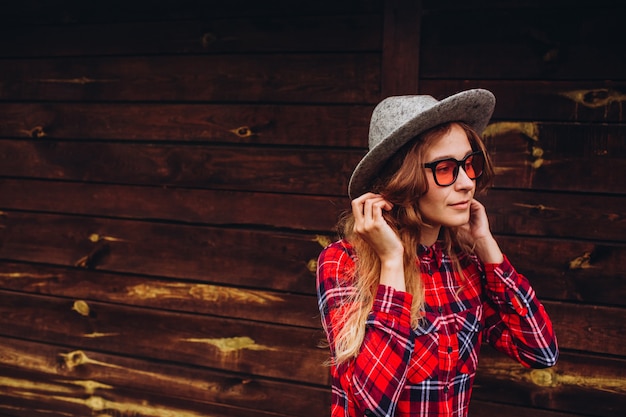 Meisje poseren tegen de achtergrond van het hek op de oude boerderij, het landelijke leven. casual meisje, gekleed in een rood en blauw geruite overhemd, jeans, zonnebril en hoed, met bruin houten hek