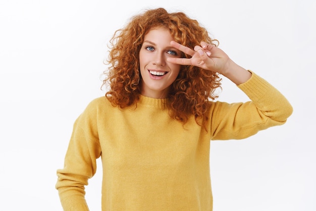 Meisje probeert positief te blijven elke situatie Aantrekkelijke gelukkige roodharige krullende vrouw in gele trui vrede overwinning teken tonen in de buurt van ogen en glimlachend permanent blij op witte achtergrond