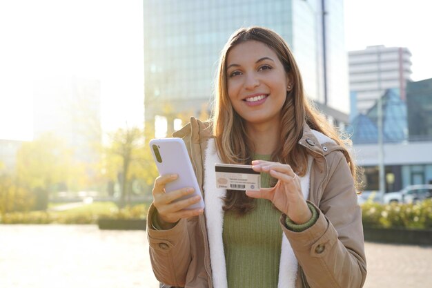 Meisje schrijft het nummer van een creditcard op zijn smartphone en doet een aankoop online terwijl hij naar de camera kijkt