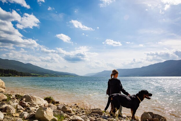 Meisje staat in de buurt van de hond van het Rottweiler-ras aan de kust in de buurt van het meer tegen de achtergrond van een bergketen bedekt met bos