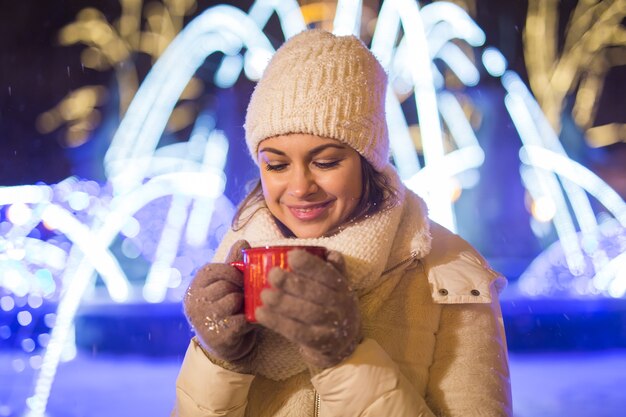 Meisje staat over winterkerststad achtergrond sneeuw sneeuw drifts, staat warme jas hoed, houdt kopje thee met warme theedranken.