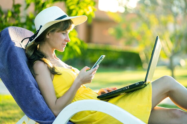 Meisje student in gele zomerjurk rusten op groen gazon in zomer park studeren op computer laptop texting op mobiele telefoon.