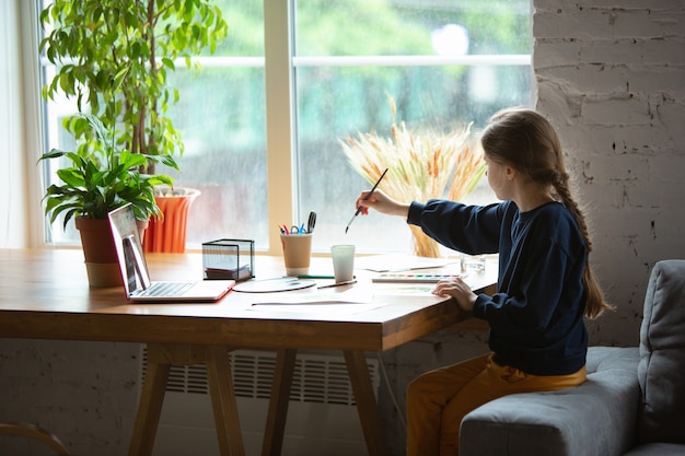 Meisje tekenen met verf en potloden thuis kijken naar docenten online tutorial op laptop