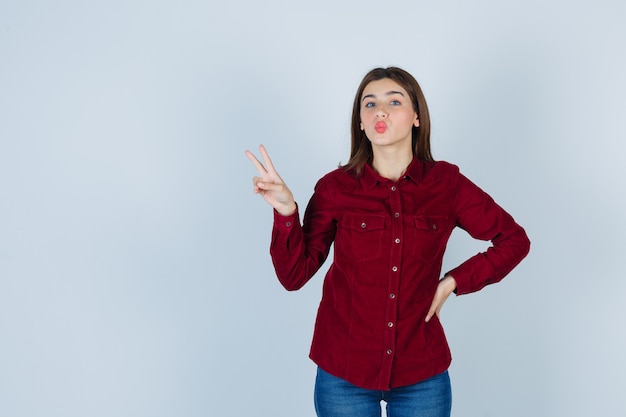 Meisje toont overwinningsgebaar, pruilende lippen in bordeauxrode blouse en ziet er positief uit