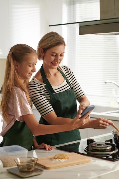 Meisje toont recept op smartphonescherm aan moeder wanneer ze saus roert terwijl ze op het fornuis kookt