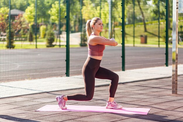 Meisje van de volledige lengte met atletisch lichaam in strakke broek training op mat buiten zomerdag beoefenen van split squats met opgeheven handen doen aerobics of pilates oefening Sportactiviteit voor gewichtsverlies