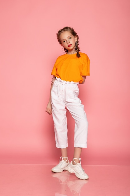 Meisje van volledige lengte op roze studiomuur. met een oranje T-shirt en een witte broek en witte sneakers.