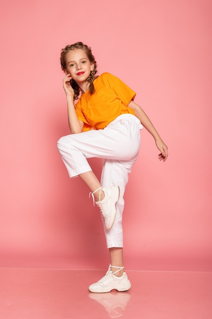 Meisje van volledige lengte op roze studiomuur. met een oranje T-shirt en een witte broek en witte sneakers.