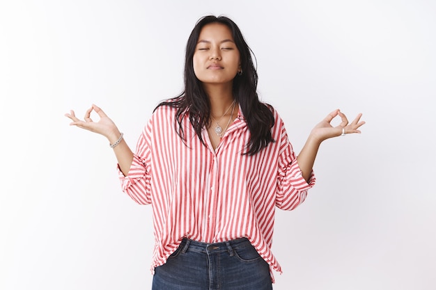 Meisje verenigt zich met natuurmeditatie met mudra-teken zijwaarts sluit de ogen en ademt lucht in, beoefent yoga in lotushouding, voelt zich ontspannen en opgelucht tegen de witte muur