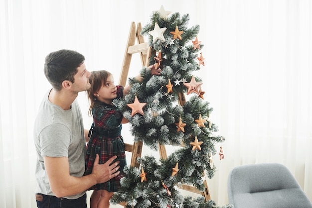 Meisje versiert mooie kerstboom met sterren terwijl haar vader haar vasthoudt.