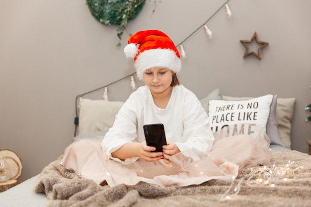 Meisje voor kerstmis, avond, tienermeisje met een kerstmuts die aan de telefoon praat terwijl ze op bed zit in een kamer die is ingericht voor kerstmis
