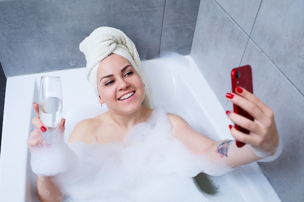 Meisje vrouw met een handdoek op haar hoofd ligt met een glas champagne in een witte badkuip en neemt een selfie aan de telefoon. In haar handen veel dronken zeep. Ontspan na een zware dag. Ontspanningsprocedure in de spa