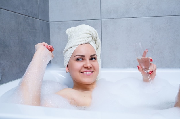 Meisje vrouw met een witte handdoek op haar hoofd ligt met een glas champagne in een witte badkuip. In haar handen veel plaszeep. Volledig whirlpoolschuim. Ontspan na een zware dag. Ontspanningsprocedure in de spa