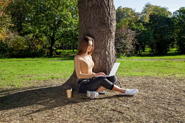 Meisje zit in de buurt van een boom in het park en werkt op een laptop Freelance werk buitenshuis