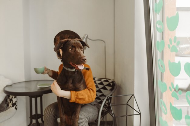 Meisje zit in een café met een kopje koffie in haar hand, de pup kijkt uit het raam