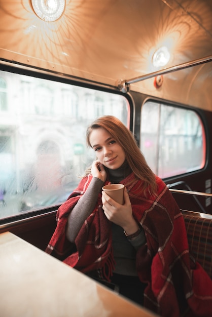 meisje zit in een coffeeshop bij het raam met een kopje koffie in haar handen, bedekt met een deken, opwarmen op een koude winterdag