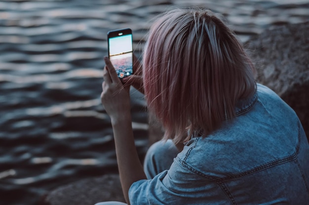 Meisje zit in een spijkerbroek en fotografeert de zonsondergang via de telefoon