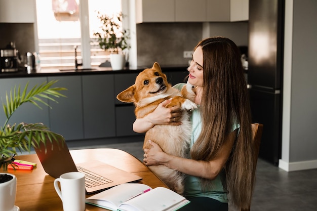 Meisje zit met laptop en knuffel Corgi-hond thuis Werken op afstand in huis Meisje speelt met mooie Welsh Corgi Pembroke in pauze op online werk Lifestyle met huisdier