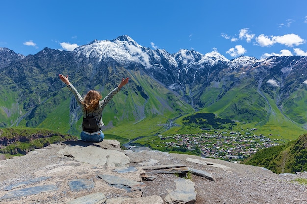 Meisje zit op de rand van de klif en kijkt naar de toppen van de bergen in de buurt van het dorp Gergeti in Georgië, onder de berg Kazbegi. Meisje kijkt naar de toppen van de bergen