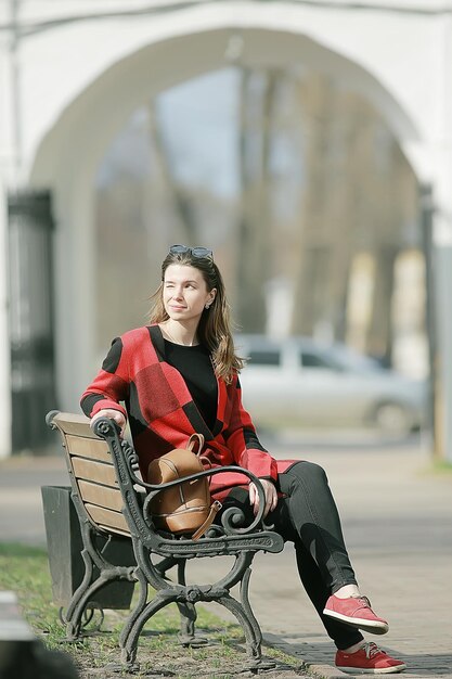 meisje zit op een tuinbank / mooi model poseren zittend in jas op bankje in stadspark, mooie meid