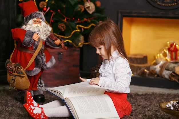 Meisje, zittend op de vloer in prachtige kerstversieringen. Meisje dat een sprookjestuk speelgoed de Kerstman leest. Meisje speelt met een stuk speelgoed Santa Claus.