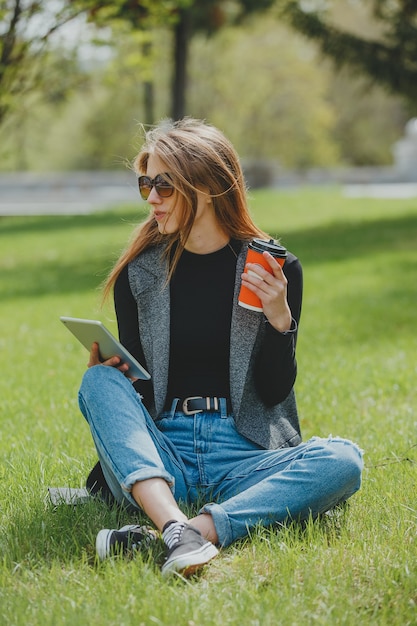 Meisje zittend op het gras met koffie en tablet op straat.