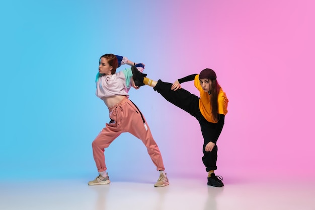 Meisjes hiphop dansen in stijlvolle kleding op gradiëntachtergrond in danszaal in neonlicht