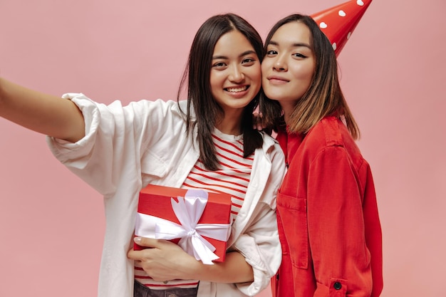 Meisjes met bruine ogen in stijlvolle oversized shirts nemen selfie-glimlach, houden rode geschenkdoos vast en vieren verjaardag op roze achtergrond