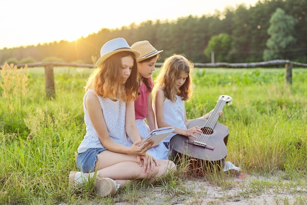 Meisjes met klassieke gitaar op aard. Kinderen ontspannen op het gazon, leren gitaar spelen, liedjes zingen, zonsondergang op de achtergrond van de zomerweide